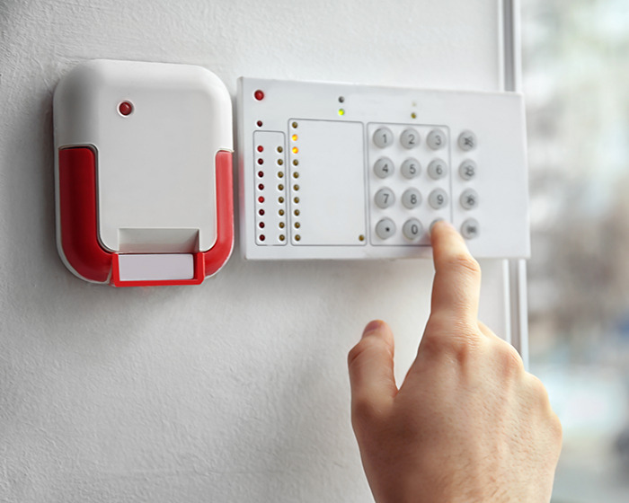Hand pressing numbers on a security alarm keypad beside a red and white wall-mounted fire alarm system.