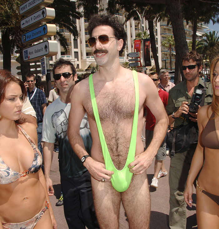 Sacha Baron Cohen in a bright green swimsuit showing his ripped transformation, surrounded by people outdoors.