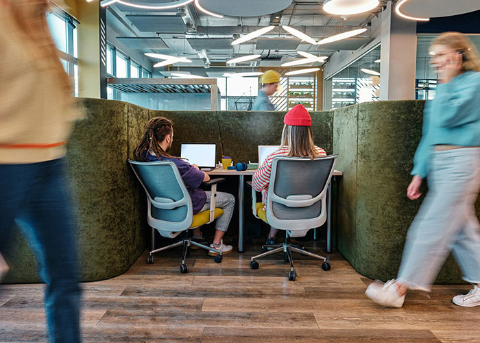 Two employees working on laptops in an office pod with blurred coworkers walking by, illustrating data fudging consequences.