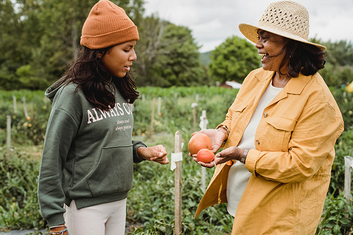 Single mom looking uncomfortable outdoors while talking to woman holding tomatoes in a garden setting.