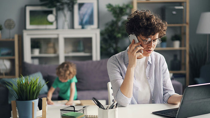 Woman talking on phone while working on laptop at home, with child in background near couch, holding phone to charge.