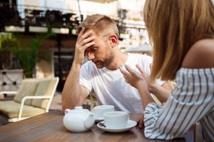 A man frustrated and holding his head while his girlfriend gestures during a tense conversation outdoors at a caf&eacute;.