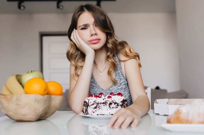 Young woman looking frustrated at cake while sitting at a table, reflecting on guy nagging her to eat more and becoming ex.
