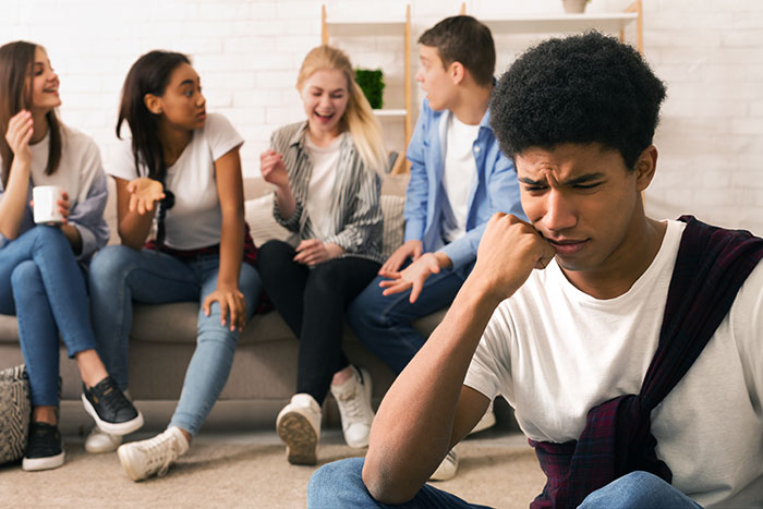 Teen boy looking upset as friends in background laugh and talk, capturing moments of realizing total jerk behavior.