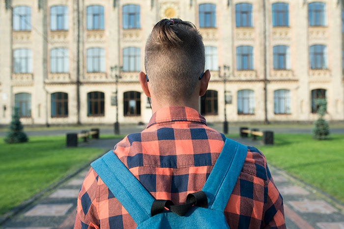 Young man with blue backpack and checkered shirt facing a large building, reflecting feelings about friends who were total jerks.