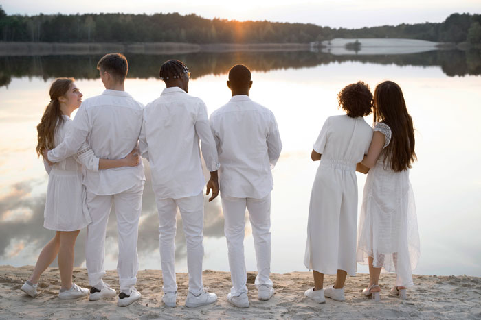 Group of diverse friends dressed in white standing by a lake at sunset ignoring the bride&rsquo;s red gown choice.