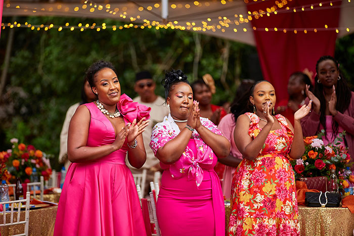 Three women at a wedding celebration, dressed in bright dresses, clapping and smiling with guests in the background.