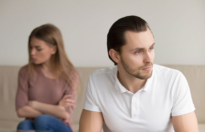 Upset man in white shirt looking away as woman sits behind him with crossed arms, highlighting secret kid and family conflict. Upset man in white shirt looking away as woman sits behind him with crossed arms, highlighting secret kid and family conflict.