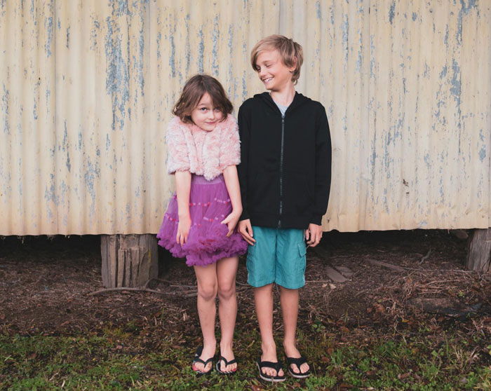 Two siblings standing outside, the golden child in purple dress looking shy while brother smiles at her.