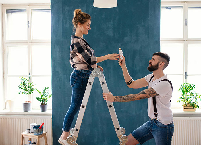 Man and woman renovating home together, sharing a moment while painting against a blue wall with natural light. - 8