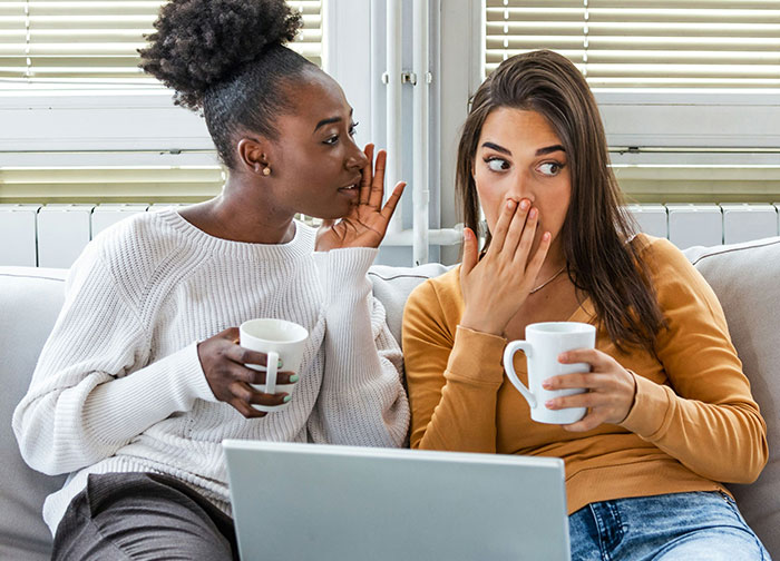 Two women sitting on a couch, one whispering a secret while holding coffee cups, illustrating honesty and emotions. - 38