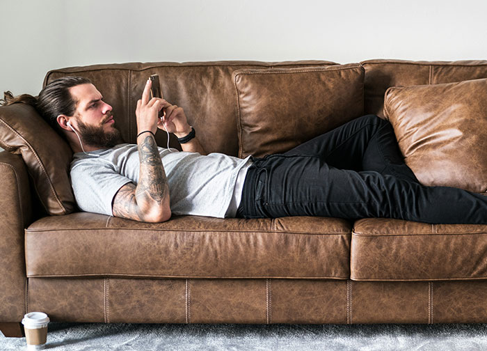 Man lying on brown leather couch using smartphone and wearing earphones, reflecting on things men never tell women. - 33