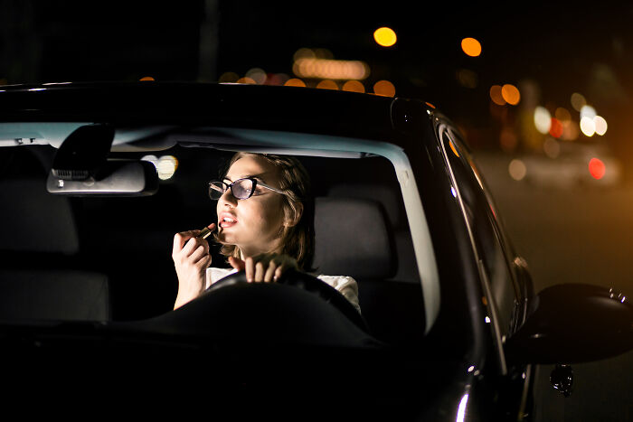 Woman wearing glasses applying lipstick while sitting in a car at night, reflecting on harmless lies parents told that caused trauma. - 16