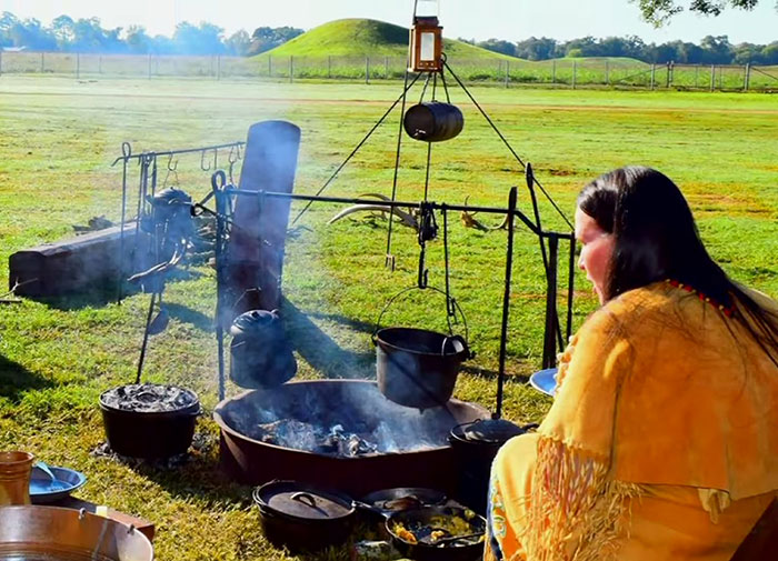 A Native American woman in traditional clothing near a fire pit demonstrating ancient language and cultural practices outdoors. - 18
