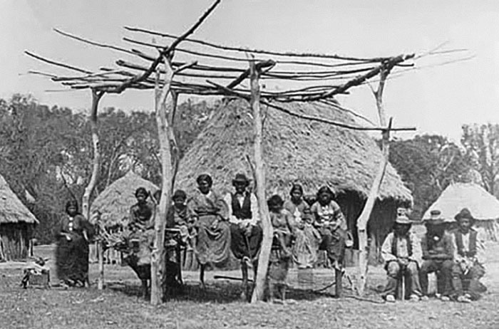 Group of Native Americans in traditional clothing sitting outside thatched huts, symbolizing language risks of extinction. - 10