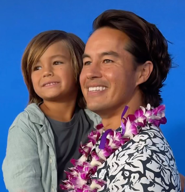Smiling dad wearing a floral lei holding his young son against a blue background, highlighting father and son relationship fears. - 2