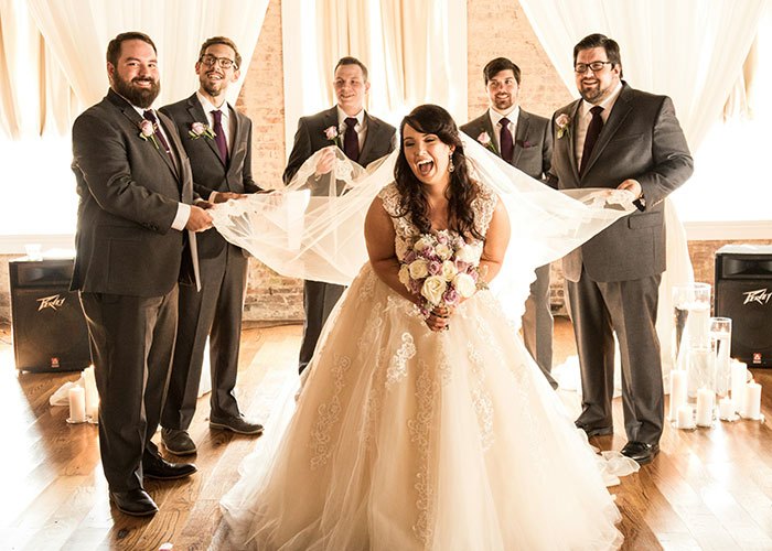 Bride laughing as groomsmen hold her veil indoors during a wedding with a call off wedding weird family tradition theme.