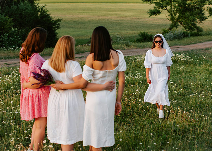 Bride in white dress and sunglasses walking toward three women in a field, illustrating call off wedding weird family tradition.