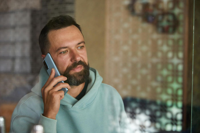 Man with beard making a phone call indoors, reflecting concern about neighbor meltdown after calling cops on 2YO wandering alone