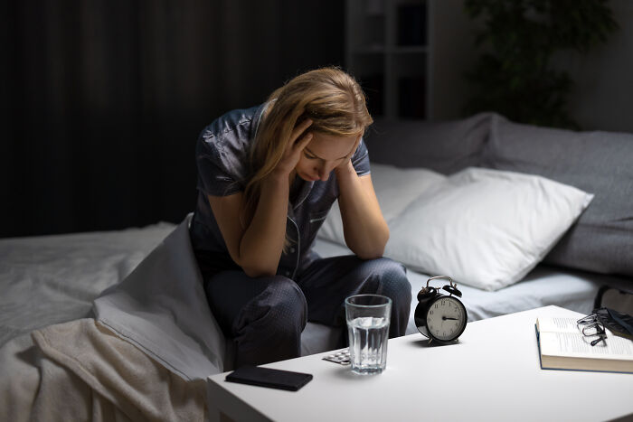 Woman sitting on bed holding head, looking distressed with pills, water glass, and alarm clock on table, cancer symptoms concept.