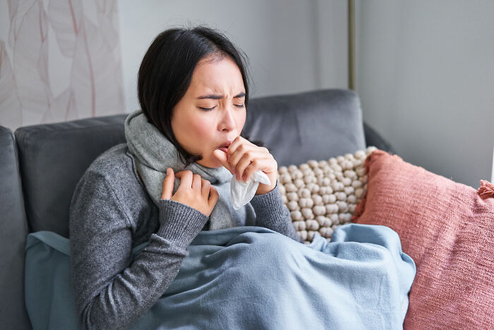 Woman wrapped in blanket coughing into hand, showing possible cancer symptoms in a home setting.