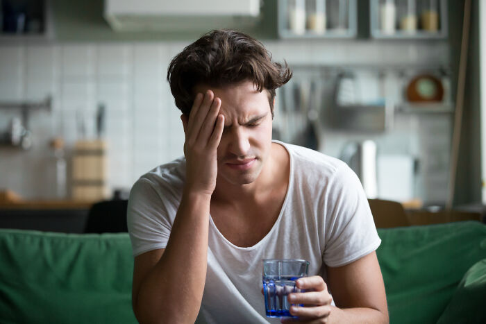 Young man sitting on a couch in pain, holding his head and a glass of water, illustrating cancer symptoms awareness.