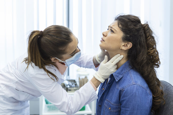 Doctor wearing gloves and mask examining a woman’s neck, focusing on cancer symptoms in a clinical setting.