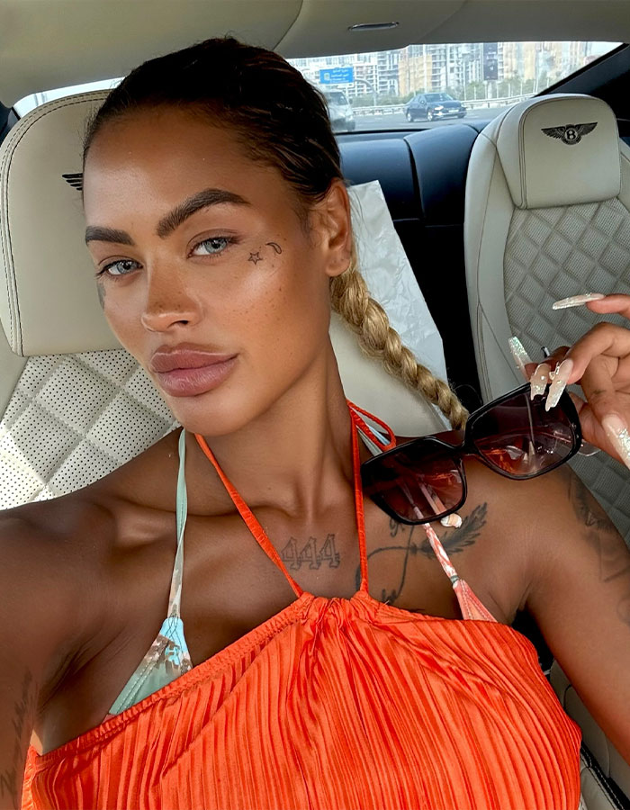 Woman with tattoos and braided hair wearing orange top and holding sunglasses inside a luxury car after butt surgery journey. - 1