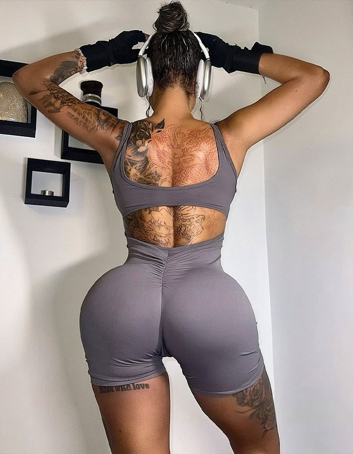 Woman wearing workout clothes and headphones, showing tattoos and enhanced butt, posing in a gym setting with gloves on. - 7