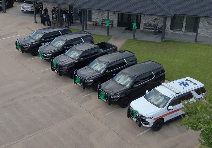 Line of black hearses and an emergency vehicle parked outside a building with people gathered nearby for a memorial event. - 13