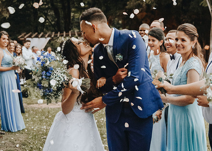 Bride and groom kissing outdoors surrounded by wedding party, capturing moment before ceremony despite caterers&rsquo; wedding date mix-up.