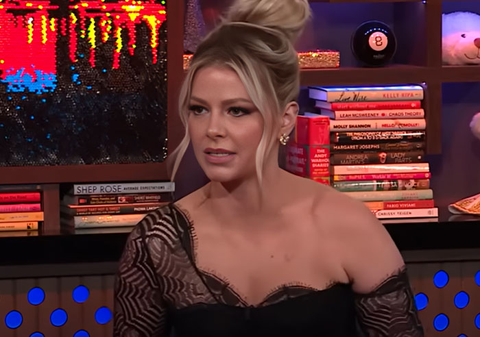 Blonde woman with hair in a bun wearing a black lace top, sitting in a colorful studio filled with books and decor items. - 30