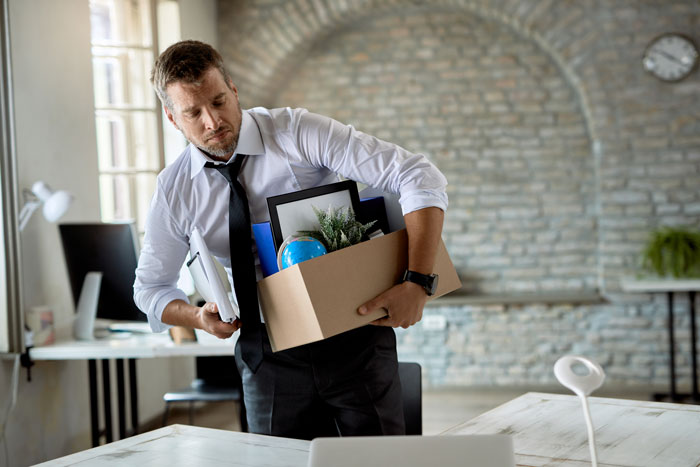 Man in white shirt and loosened tie carrying a box of office items after tracking missing equipment for months.
