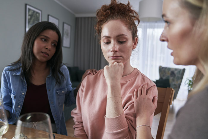 Three women indoors, one comforting a distressed young woman, reflecting concern in a supportive home setting.