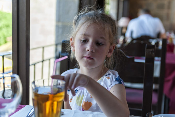 8YO girl sitting at a table with a drink, looking curious as she points, interacting with strangers nearby.