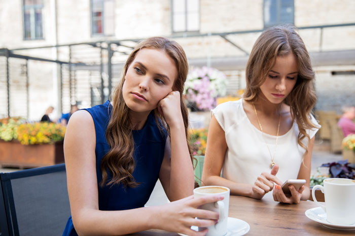 Two women sitting at an outdoor caf&eacute;, one looking concerned while the other focuses on her phone, mom and friend scene.