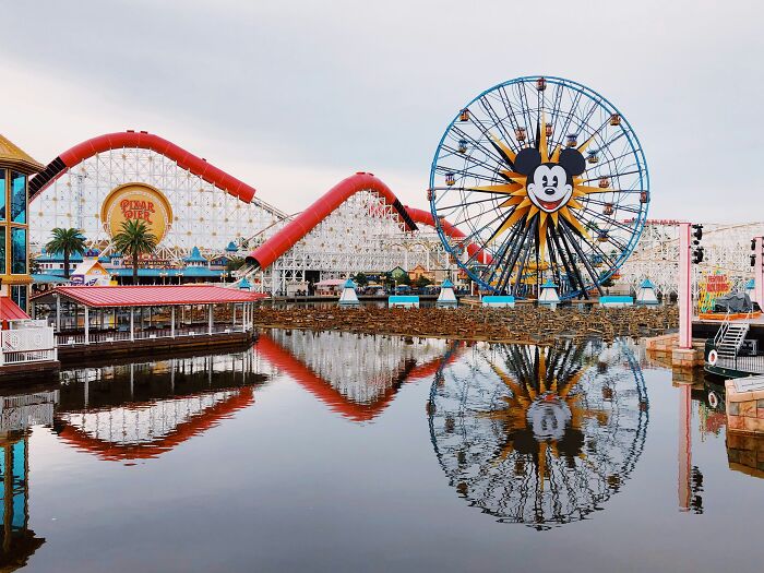 Amusement park with a large Ferris wheel featuring a cartoon face, roller coaster, and water reflections on a calm day. - 14