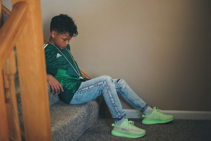 Boy sitting on carpeted stairs looking down, wearing green shirt and bright green sneakers, evoking disturbing things overheard.