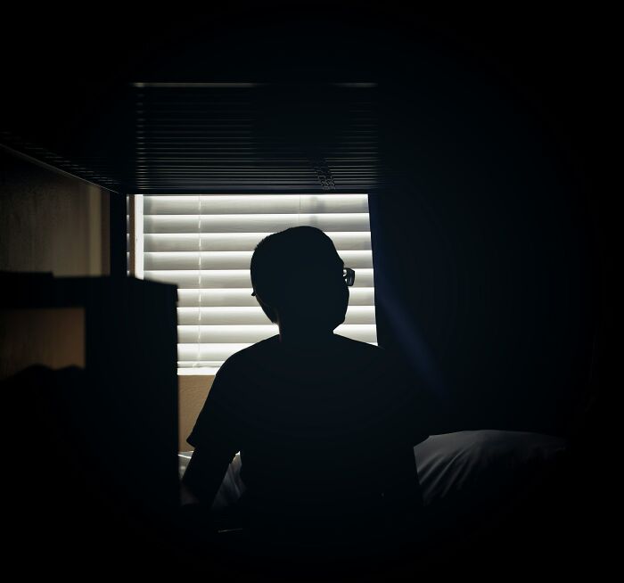 Silhouetted child sitting alone in dark room near window blinds, illustrating severe child punishment and neglect.