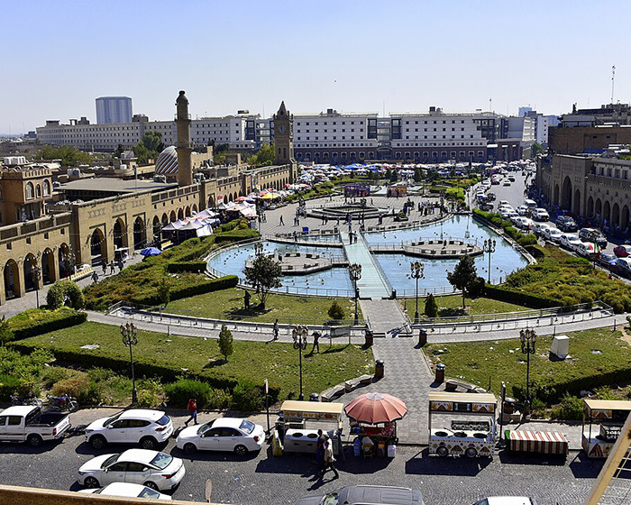 Wide view of a city park with fountains, historic buildings, and cars parked around, highlighting city bans and dark conspiracy concerns.
