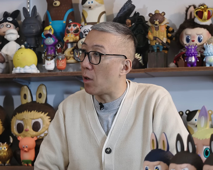 Man in glasses and beige sweater speaking indoors, surrounded by Labubu and other collectible figures on shelves.