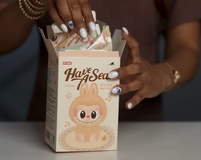 Hands with white nail polish opening a Labubu product box, highlighting City bans Labubu dark conspiracy concerns.