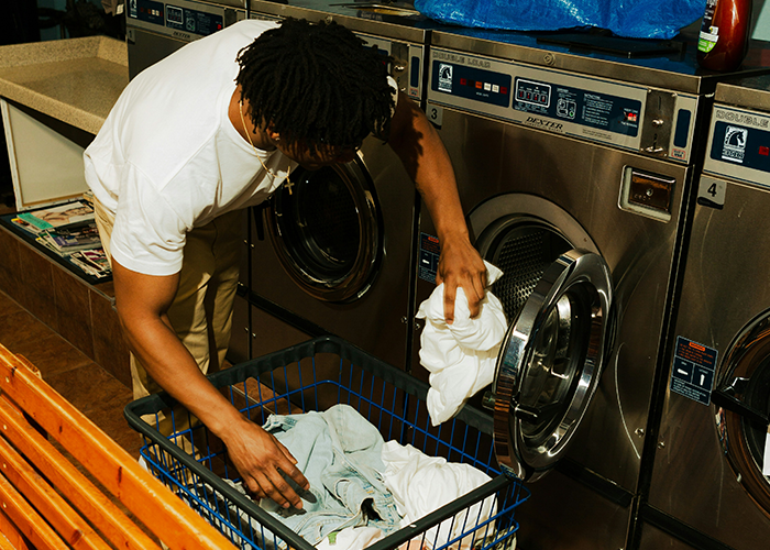 College student loading laundry in a washing machine at a laundromat, showing responsibility and cleaning efforts. - 25