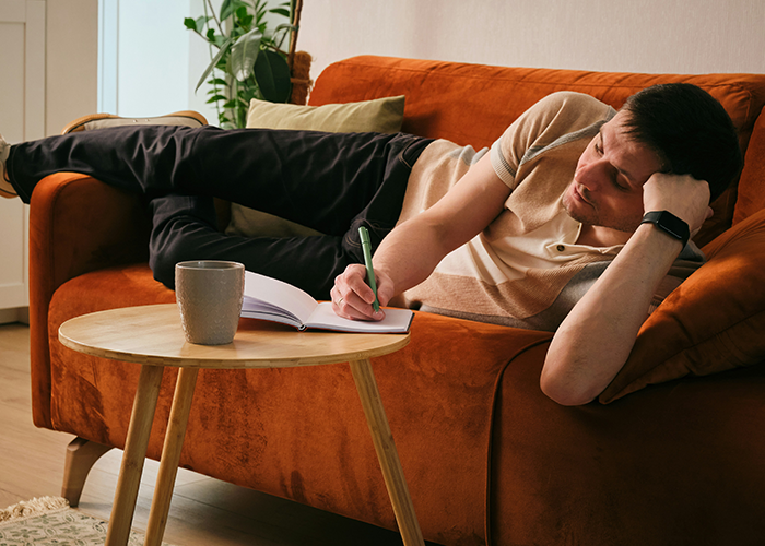 College student writing in notebook while lounging on orange couch in tidy shared apartment with plant in background. - 26