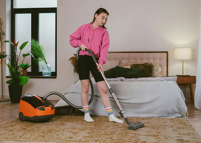 College student vacuuming bedroom carpet, wearing pink hoodie and striped socks, showing tidy roommate behavior. - 18