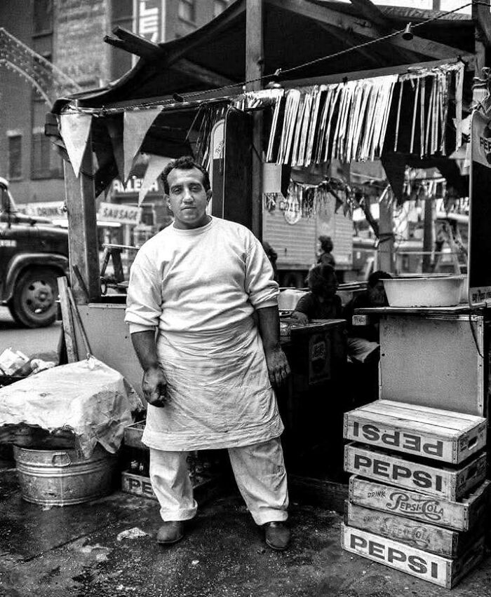 Black and white photo of a street vendor standing by a food stall with vintage Pepsi crates, unique old photos perspective.
