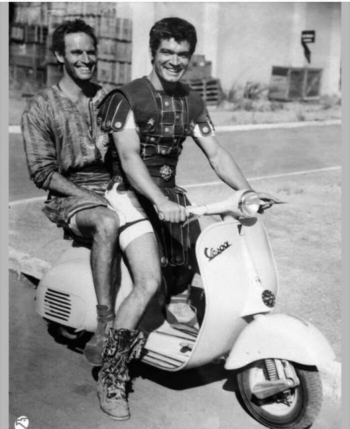 Two men in vintage costumes smiling while riding a classic Vespa scooter in a unique old photo.