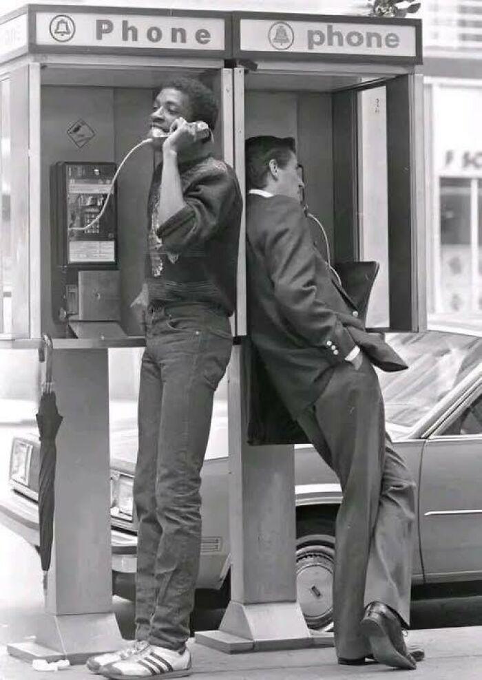 Two men using adjacent phone booths, one smiling and the other leaning back, unique old photos capturing moments.