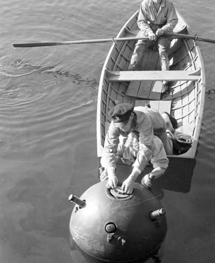 Two men in a rowboat handling an old naval sea mine in calm water, a unique old photo with an incredible story behind it.