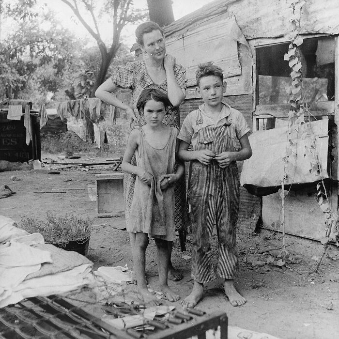 Three children standing barefoot outside a rustic wooden shack in a unique old photo from the Great Depression era.